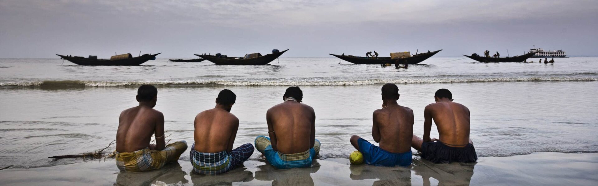 A row of men seated on the wet sand at low tide, praying for a benevolent sea, before going fishing, after one of the strongest cyclones to hit the country killed thousands of fishermen at sea.